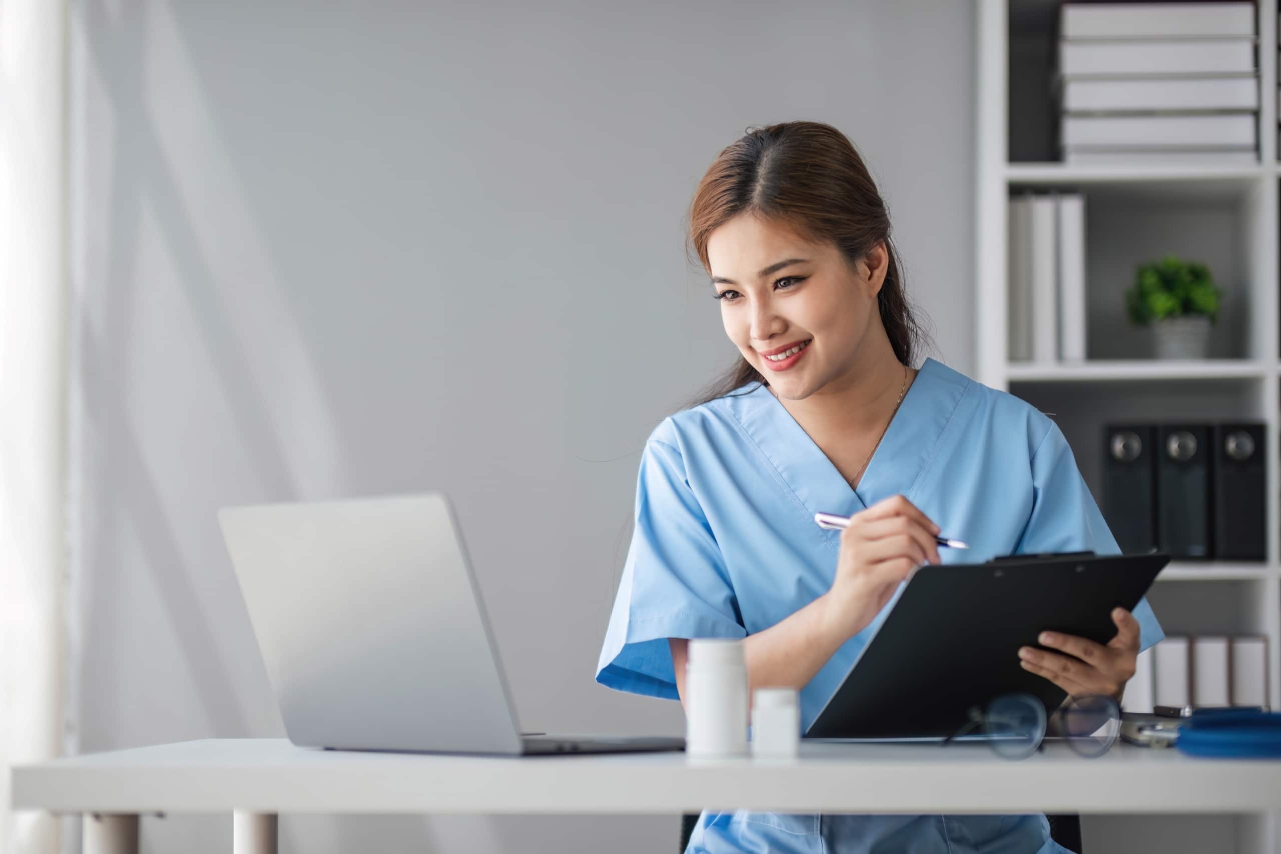 Asian medical professional using a laptop and writing on a clipboard